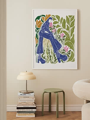 Stampa artistica colorata incorniciata con figura seduta tra grandi foglie e fiori su parete beige