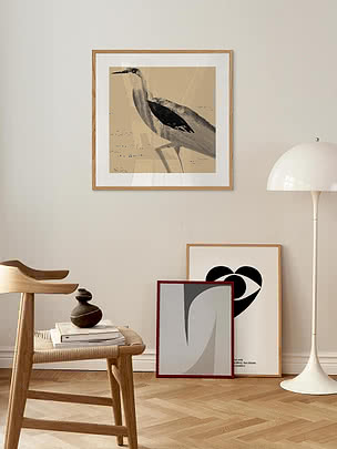 Stampa Heron in cornice di rovere con passepartout bianco su parete chiara, sopra una sedia in rovere e una panca di libri