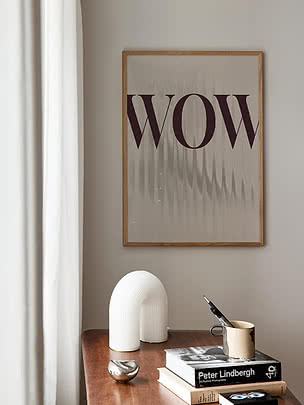 Poster WOW in eiken lijst op een greige wand boven een notenhouten dressoir met witte booglamp, keramieken mok en fotoboeken