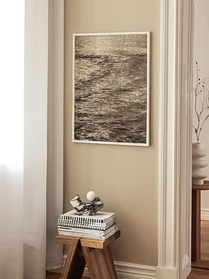 Print Warm Tide in lichte lijst op een beige muur in een smalle nis boven een houten kruk met boeken en een sculptuur