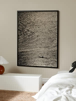Print Low Tide in zwarte lijst op een beige slaapkamermuur boven een witte bank, met een bed in wit linnen ernaast