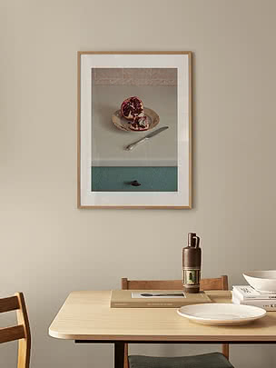 Split Fruit poster in an oak frame on a beige wall above a light oak dining table with plates and a ceramic vase