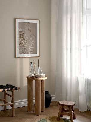 Dried Bloom poster in an oak frame on a beige wall above a round wooden side table with brushes and a black vase