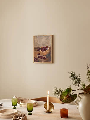 Red Cherries i egeramme på beige væg over et dækket bord med lærred, cremefarvet stentøj, riflede grønne glas og en messinglysestage formet som en musling