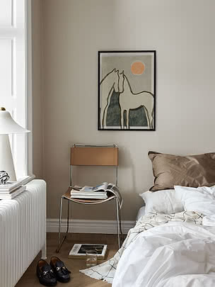 Poster Equine Embrace im schwarzen Rahmen an beiger Schlafzimmerwand über einem cognacfarbenen Lederstuhl mit Zeitschriften, neben einem Bett in weißem Leinen