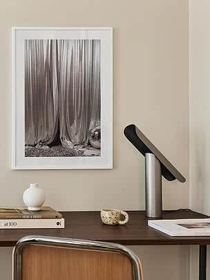 Drape in a white frame with a wide mat on a beige wall above a dark wood desk with a brushed aluminium lamp, book stacks and an open magazine