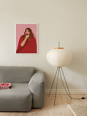 Plakaten Cozy Woman Apple i hvid ramme på en cremefarvet væg over en grå hørsofa ved siden af en gulvlampe i papir