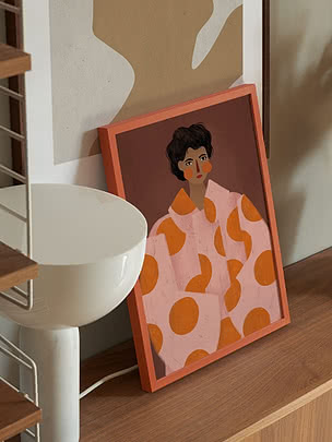 Print Fashion Portrait in koraalkleurige lijst op een notenhouten dressoir naast een witte koepellamp en een draadwandrek