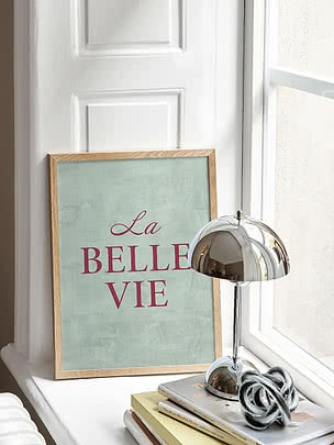 Stampa La Belle Vie in cornice di rovere appoggiata su un davanzale accanto a una lampada cromata e una pila di libri
