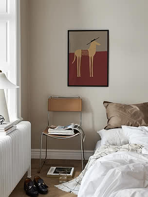 Poster Horse with Sombrero in cornice nera su parete beige di camera sopra una sedia in pelle carica di riviste, accanto a un letto rifatto in lino bianco
