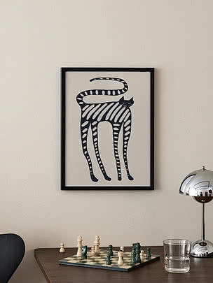 Affiche Striped Whimsical Cat dans un cadre noir sur un mur crème au-dessus d'un bureau en bois sombre avec un échiquier en pleine partie, un verre d'eau et une lampe de table chromée