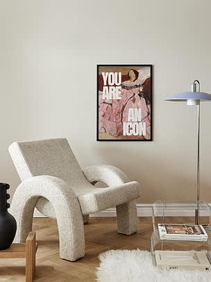 Poster You are an Icon in cornice nera su parete bianco sporco sopra una poltrona in bouclé crema, accanto a una piantana e un portariviste in filo
