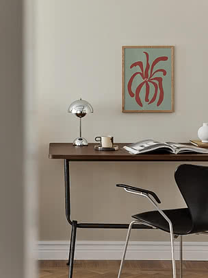 Muted Organic Bloom poster in a light oak frame on a pale wall above a dark wood desk with a chrome mushroom lamp, an espresso cup and an open magazine, beside a black chair