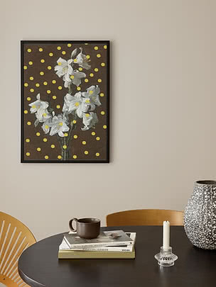 Plakaten Lilies Polka Dots i svart ramme på en beige vegg over et rundt mørkt spisebord med et krus i steingods, bøker, en glasslysestake og en spettet vase