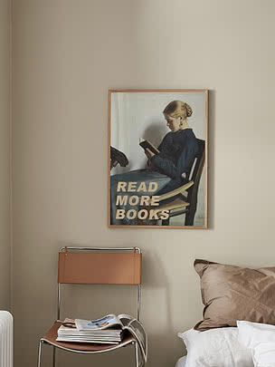 Plakaten Woman Reading Book i eikeramme på en kremfarget soveromsvegg over en stol i cognacfarget skinn og krom med en stabel magasiner, ved siden av en seng med en støvet rosa pute