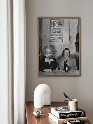 Plakaten Retro Disco Bar i eikeramme på en beige vegg over en konsoll i mørkt tre med en riflet hvit lampe, et kremfarget krus med penner og en fotobok