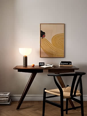 Woman Golden Waves poster in an oak frame on a pale wall above a dark wood desk with a glass table lamp, an open notebook and a black wishbone chair
