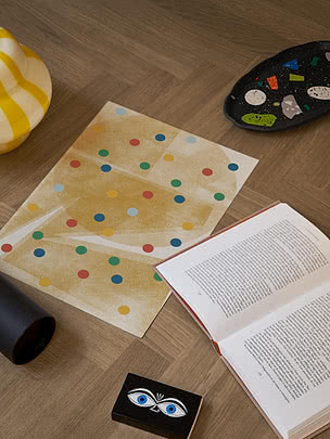 Plakat Polka Dot Harmony leżący bez ramy na podłodze w jodełkę obok otwartej książki, żółto-białego wazonu w paski, czarnej cętkowanej miseczki i małego zadrukowanego pudełka