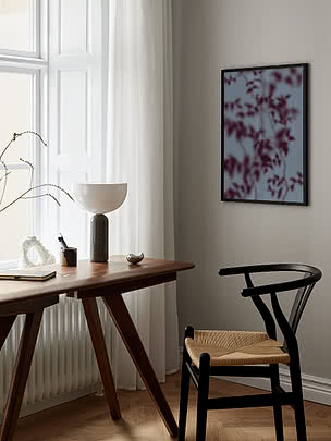 Botanical Shadows I poster in a black frame on a greige wall beside a dark wood desk with a white hemisphere lamp, a book and a brass dish, next to a black wishbone chair