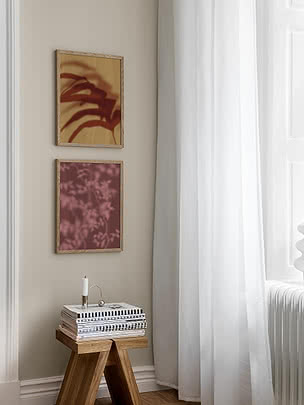 Botanical Shadows II poster in an oak frame hung below a second botanical print on a beige wall, beside a white curtain and a wooden stool with striped books and a candleholder