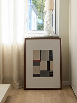 Plakat Geometric Harmony w ramie z ciemnego drewna oparty o ścianę pod oknem z lnianą zasłoną, obok biała fakturowana lampa na parapecie, na podłodze z jasnego drewna