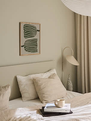 Abstract Tulips poster in an oak frame on a greige bedroom wall above a beige headboard with corduroy cushions, beside a cream wall lamp and a speckled vase on books