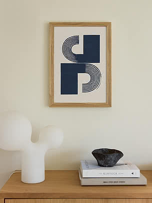Plakat Modern Objects Blue w dębowej ramie na kremowej ścianie nad dębową komodą z białą rzeźbiarską lampą, stosem książek i misą z ciemnego kamienia