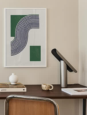 Plakat Modern Green Lines w białej ramie na kremowej ścianie nad orzechowym biurkiem z czarną uchylną lampą, okrągłym białym wazonem, książkami i cętkowanym kubkiem, obok krzesło z plecionką