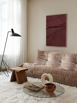 Plakaten Oxblood Plume i egeramme på en cremefarvet væg over en beige sofa i riflet fløjl, ved siden af en sort vinklet gulvlampe og en træskammel, med et glasbord på et loddent hvidt tæppe