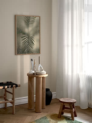 Frosted Palm I poster in an oak frame on a greige wall beside a white curtain, above a round wooden table with ceramics and brushes, with a black vase and a wooden stool below