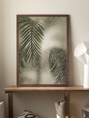 Frosted Palm II poster in a dark wood frame standing on a wooden table against a cream wall, beside a white paper lamp, with paper rolls and a patterned vase below