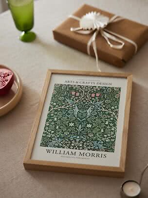 Framed William Morris print on a table beside a wrapped gift, green glass and pomegranate