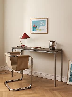 Hemmkontor med smalt skrivbord, röd bordslampa, stålkanna och stol med sittdel i canvas