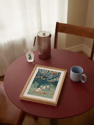 Framed Moomin print on a red table with a brown pitcher, glass, and blue mug