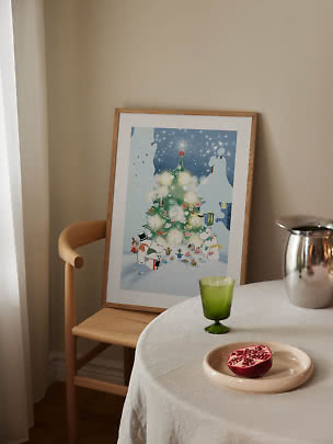 Affiche de Noël Moomin encadrée sur une chaise en bois, près d’une table avec verre vert.