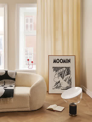 Soggiorno luminoso con poltrona color crema, tenda gialla e poster Moomin appoggiato al muro.