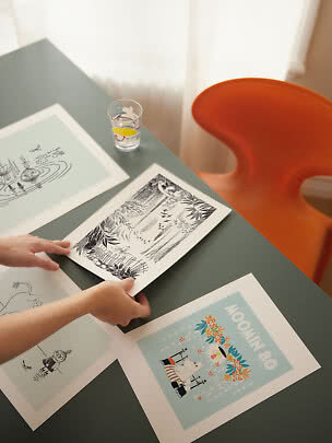 Mani sistemano stampe illustrate su un tavolo scuro accanto a una sedia arancione