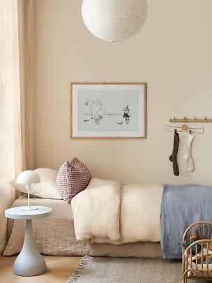 Cameretta accogliente con letto basso, cuscini, piccola lampada bianca e quadro incorniciato.