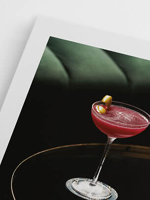 Cocktail rosso in coppa con scorza di limone su vassoio nero davanti a una stampa incorniciata