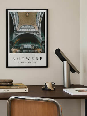 Framed Antwerp Central Station poster above a desk with lamp, books, and cup