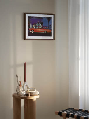 Framed retro car print on a light wall above a round wooden table with candle holder and decor
