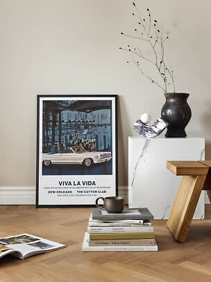 Framed vintage poster leaning against wall beside vase, books and wooden stool