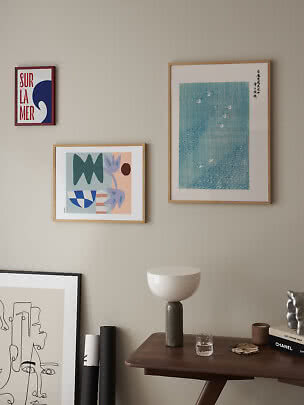 Tre indrammede kunstplakater på beige væg over et mørkt træbord med lampe og glas.