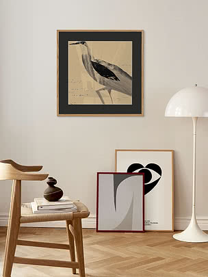Poster con airone in passepartout nero e cornice in rovere su una parete beige, sopra uno sgabello di legno e una piantana