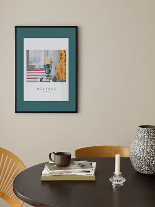 Framed Matisse poster with teal mat above a round table with books, mug and candlestick