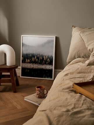 Soveværelse med beige sengetøj, naturplakat på gulvet og hvid lampe på træskammel