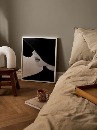 Framed black-and-white poster of a woman in a hat, leaning by a bed in a warm-toned bedroom