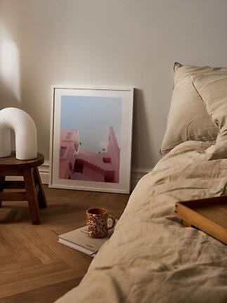 Mysigt sovrum med beige sängkläder, vit bordslampa på pall och inramad rosa konstbild