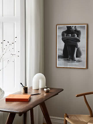 Zwart-wit poster in houten lijst boven een bureau met lamp, boek en stoel bij een raam