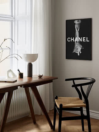 Stilren arbetsplats med träskrivbord, svart stol, bordslampa och inramad Chanel-affisch på väggen